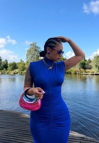 Robe bleue ajustée avec une texture froncée, manches courtes et col haut ; tenant un sac à main brillant en rose et métallique, accessoirisé avec des lunettes de soleil.