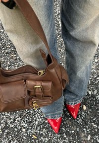 Καφέ δερμάτινη τσάντα χειρός με τσέπη με φερμουάρ και λεπτομέρεια από αγκράφα, συνδυασμένη με μπλε τζιν και γυαλιστερά κόκκινα ψηλοτάκουνα παπούτσια σε gravel.