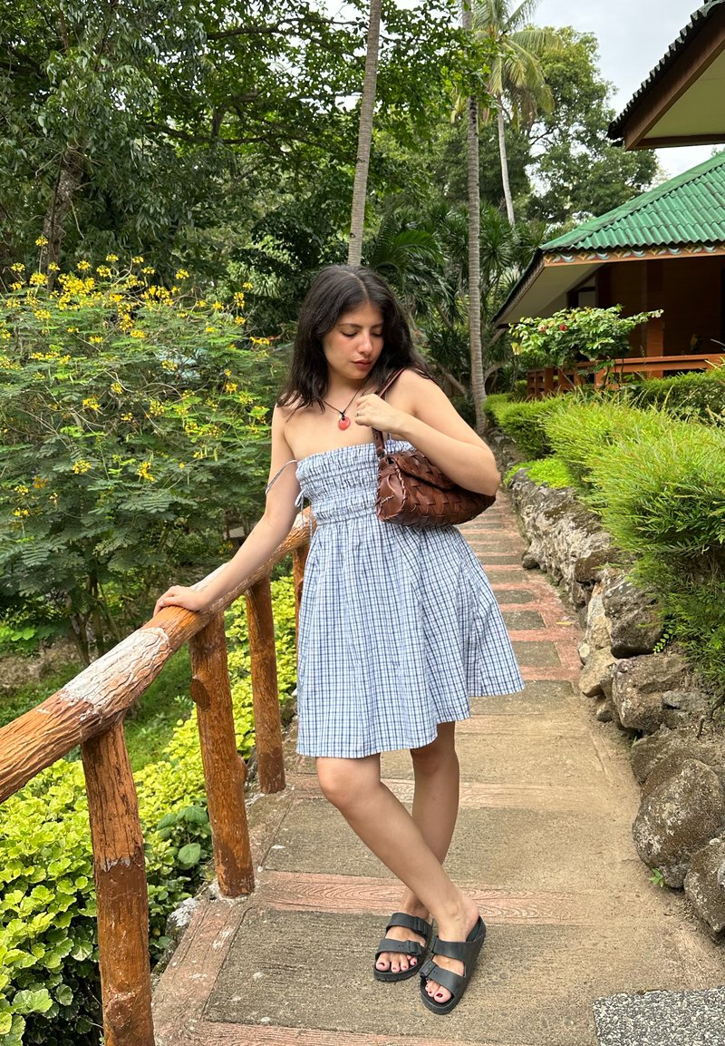 Mladá žena v modrých károvaných šatech bez ramínek a černých sandálech stojí na zahradní cestě, drží dřevěné zábradlí a hnědou pletenou tašku, obklopena zelení.