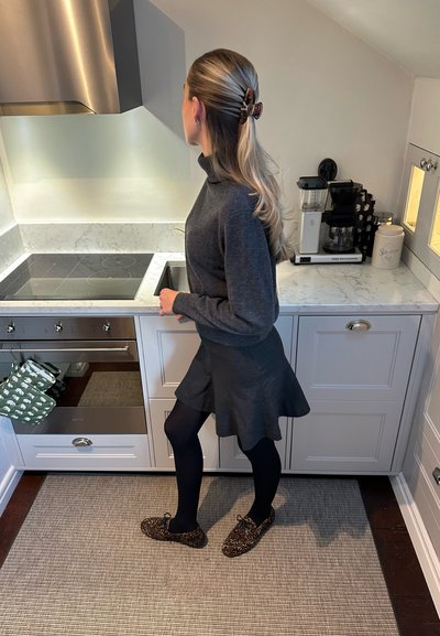Suéter de cuello alto gris oscuro, falda acampanada, medias negras y zapatillas de leopardo; cocina moderna con encimera de mármol y electrodomésticos de acero inoxidable.