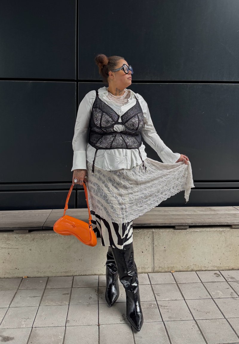 Woman posing against black wall wearing white blouse with black lace bustier, patterned skirt, black boots, round sunglasses, and holding orange handbag.
