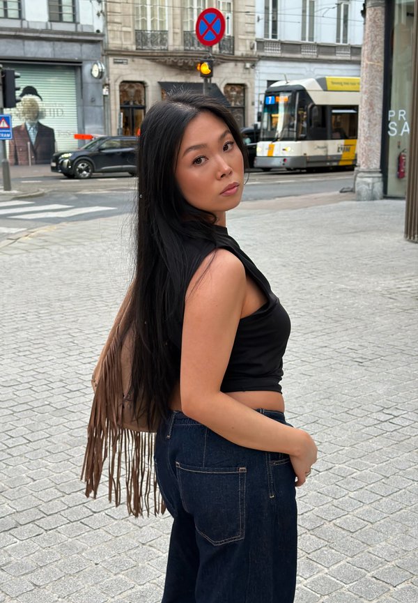 Femme aux longs cheveux noirs portant un haut noir sans manches et un jean, se tient dans une rue de la ville près d'un tramway et d'un feu de circulation jaune.