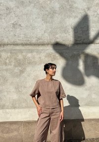 Brązowa lniana bluzka z krótkimi rękawami w połączeniu z szerokimi lnianymi spodniami. Miękka tekstura, minimalistyczny design, na tle teksturowanej ściany.