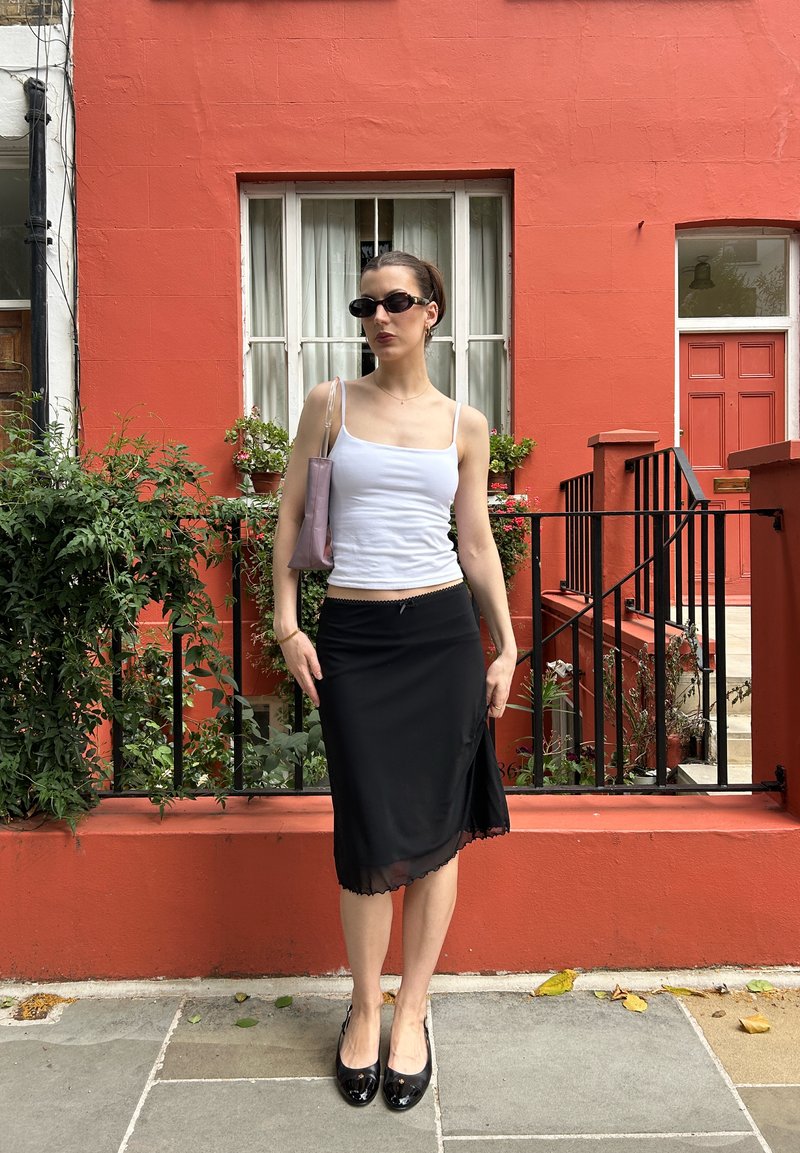 Femme portant une jupe noire, un débardeur blanc, des lunettes de soleil noires et des chaussures noires, debout devant un bâtiment rouge avec une fenêtre et une porte.