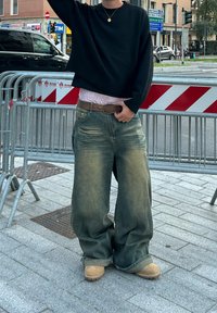 Større lyseblå denimjeans med slidte detaljer, parret med en cropped sort sweater. Bæreren står på en stenbelægning.