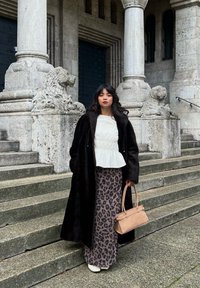Svart pelsjakke av kunstpels over hvit peplum-topp og leopardmønstret maxiskjørt. Beige håndveske og sko med hæl. Steintrapper og søyler i bakgrunnen.