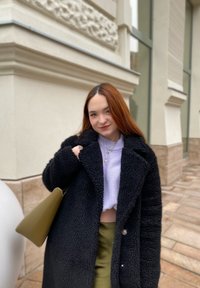 Čierny nadýchaný kabát cez svetlo fialový sveter a zelenú suk�ňu, drží khaki tašku. Prejavuje vrstvené náhrdelníky a dlhé, rovné vlasy.