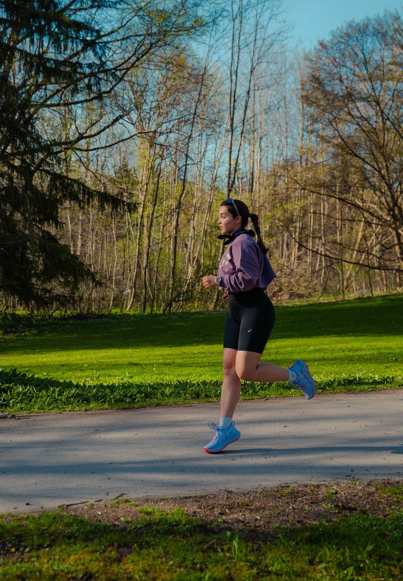 Femme faisant du jogging sur un chemin pavé dans un parc avec de l'herbe verte et des arbres sans feuilles sous un ciel bleu clair.