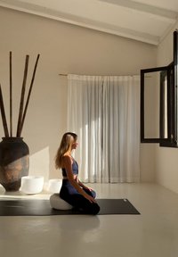 Mata do jogi w jasnym pomieszczeniu z gładką, białą poduszką. Duży czarny wazon z łodygami bambusa, delikatne białe zasłony i promienie słoneczne oświetlają przestrzeń.