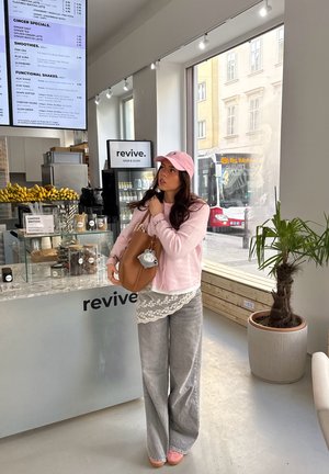 Joven mujer con gorra y suéter rosa sosteniendo una bolsa beige dentro de un bar de jugos luminoso llamado Revive, con plátanos y una pizarra de menú.