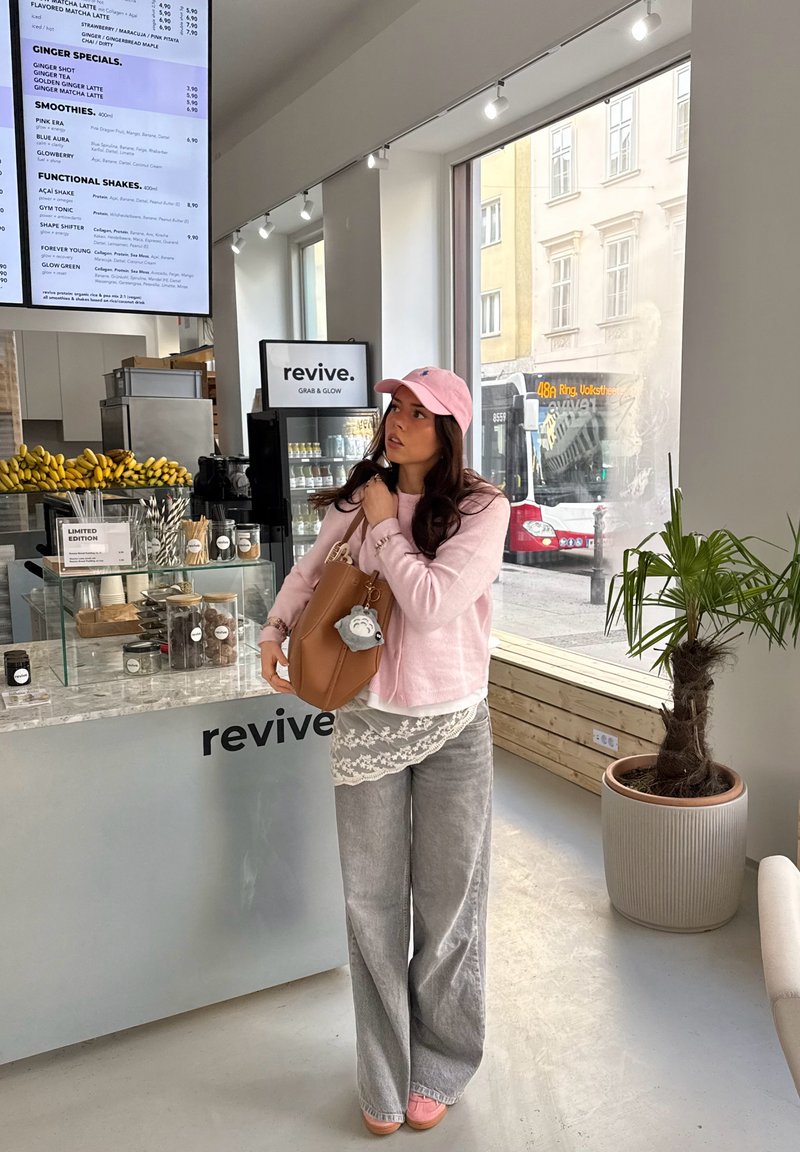 Jeune femme portant une casquette rose et un pull, tenant un sac beige à l'intérieur d'un bar à jus lumineux nommé Revive, avec des bananes et un tableau de menu.