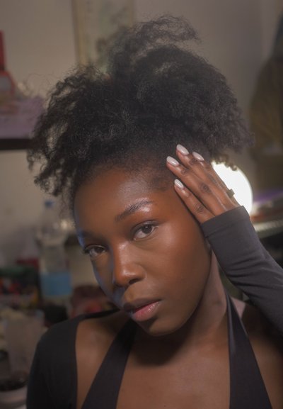 Joven con cabello rizado natural, tocándose la cabeza, vestida con ropa negra, en un ambiente interior iluminado suavemente con objetos de fondo desenfocados.