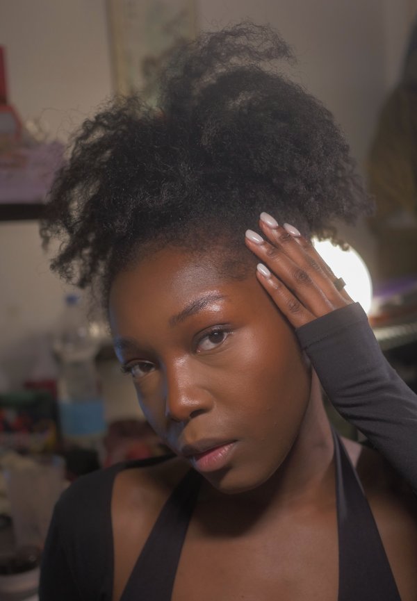 Giovane donna con capelli ricci naturali, che si tocca la testa, vestita di nero, in un ambiente interno illuminato dolcemente con oggetti sullo sfondo sfocati.