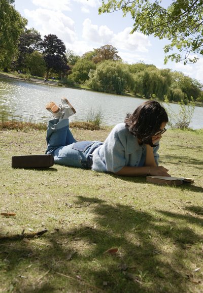 Una persona yace sobre la hierba junto a un lago, vestida con un suéter difuso azul claro y jeans de denim, con botas marrones. Una bolsa marrón descansa cerca.