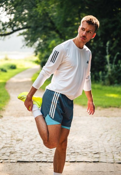 Camisa deportiva de manga larga blanca con tres rayas negras. Pantalones cortos grises y verde azulado, zapatillas deportivas de color amarillo neón, estirándose en un camino.