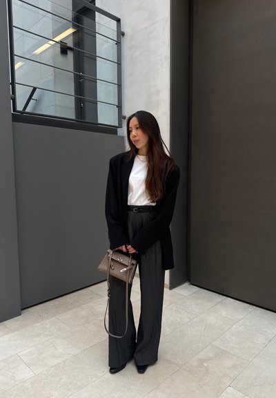 Blazer negro sobre una camiseta blanca, pantalones de tiro alto a rayas verticales y zapatos de punta negros. Sosteniendo un bolso de mano marrón claro con herrajes plateados.