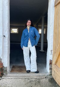 Niebieska koszula dżinsowa z przednią kieszonką, zestawiona z białymi spodniami o szerokich nogawkach. Noszona z lekkim szalem, z teksturowanym dywanem przy wejściu.