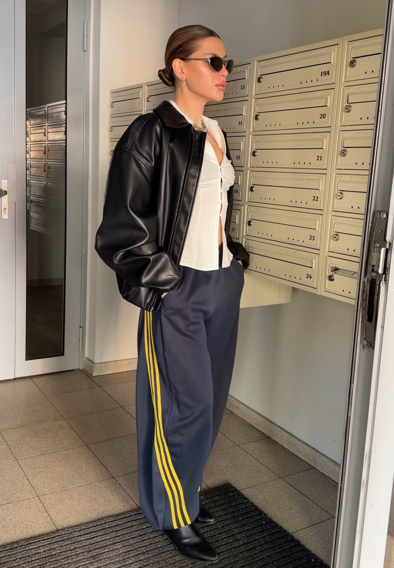 Γυναίκα με γυαλιά ηλίου, φορώντας μαύρο δερμάτινο μπουφάν, λευκό πουκάμισο και ναυτικό μπλε παντελόνι με κίτρινες ρίγες, στέκεται δίπλα σε μπεζ θυρίδες ταχυδρομείου σε εσωτερικό χώρο.