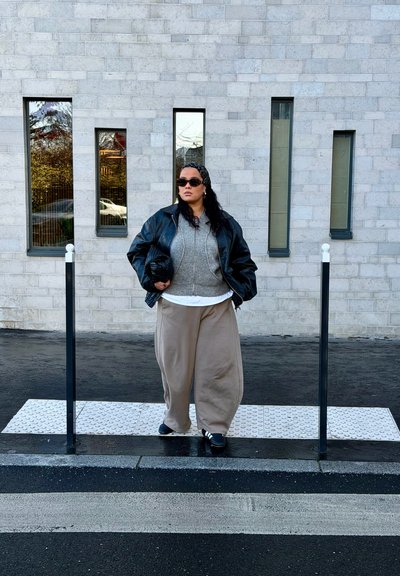 Chaqueta de cuero negra, suéter gris con cremallera, camisa blanca, pantalones beige holgados y zapatos negros. De pie sobre un pavimento táctil cerca de una pared gris.