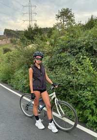 Crna majica bez rukava, crne rukavice, bijele tenisice; stoji pored bijelog planinskog bicikla na teksturiranoj cesti s zelenilom u pozadini.