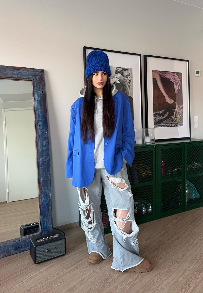 Γυναίκα που στέκεται μέσα σε εσωτερικό χώρο φορώντας μπλε σκουφάκι, μπλε σακάκι, γκρι φούτερ με κουκούλα, γκρι παντελόνι με έντονες σκισίματα και καφέ παντόφλες κοντά σε καθρέφτη και κορνιζαρισμένη τέχνη.