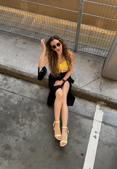Top corto amarillo, falda negra y sandalias de tacón alto amarillas. Cabello largo y rizado, y gafas de sol oscuras. Fondo de concreto con una valla.