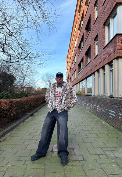 Chaqueta de piel sintética con estampado de leopardo marrón, suéter ligero con texto, jeans de pierna ancha oscuros, zapatillas negras, de pie en un camino pavimentado junto a la vegetación.