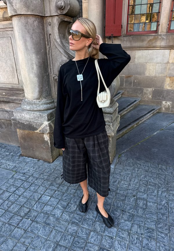 Femme blonde portant des lunettes de soleil surdimensionnées, un haut à manches longues noir, un short à jambes larges à carreaux, des chaussures noires et un sac à épaule blanc, debout près des marches en pierre d'un bâtiment.