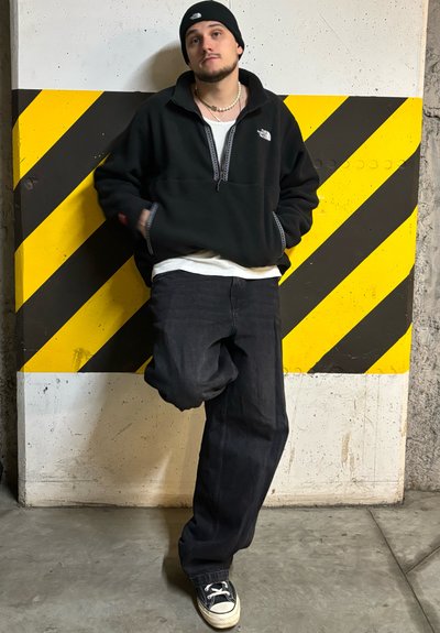 Sudadera de forro polar negra con cuello con cremallera, jeans oscuros y zapatillas negras. El fondo presenta una pared con rayas amarillas y negras.