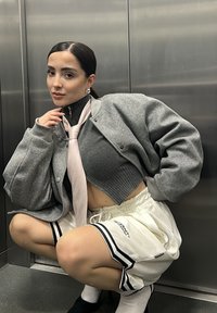 Šedá oversized bomber bunda přes tmavý žebrovaný crop top, doplněná bílými sportovními šortkami s černými pruhy a textem. Doplňky zahrnují růžový náhrdelník.