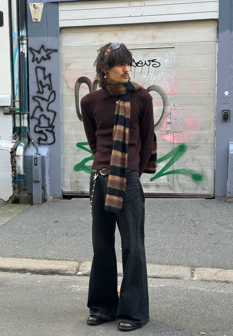 Pantalon noir évasé associé à un pull marron côtelé et une écharpe rayée marron/noir. Des bottes en cuir complètent le look. En arrière-plan, un graffiti.