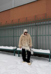 Personne portant une veste beige duveteuse, un pantalon noir, un haut crème orné de dentelle et des lunettes de soleil, debout sur la neige devant une clôture en métal verte et des bancs.