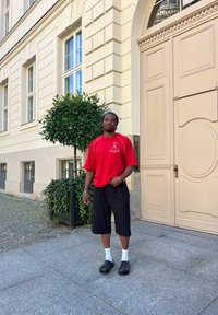 Κόκκινο κοντομάνικο πουκάμισο με λογότυπο, μαύρη βερμούδα μέχρι το γόνατο, λευκές κάλτσες και μαύρα εσπαντρίγια. Το φόντο έχει μπεζ κτίριο με παράθυρα.