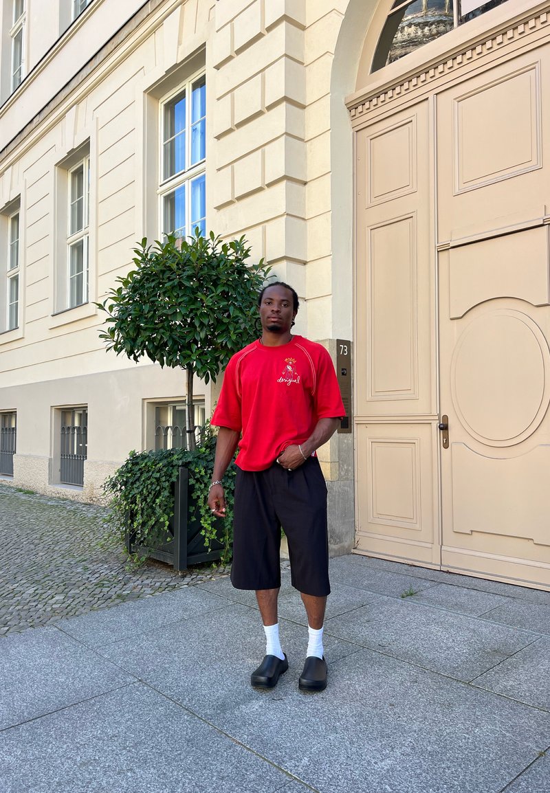 Κόκκινο κοντομάνικο πουκάμισο με λογότυπο, μαύρη βερμούδα μέχρι το γόνατο, λευκές κάλτσες και μαύρα εσπαντρίγια. Το φόντο έχει μπεζ κτίριο με παράθυρα.