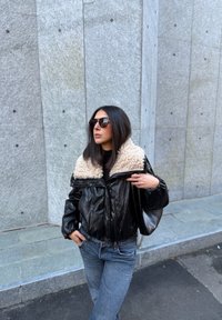 Veste courte en cuir noir avec un col en fausse fourrure beige ; associée à un jean bleu clair. Des lunettes de soleil noires complètent le look.