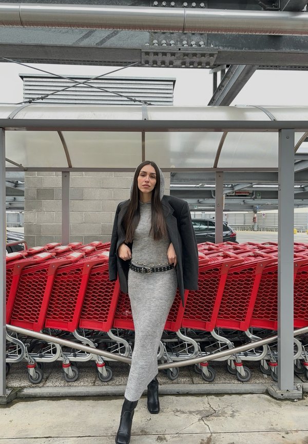 Vestido gris de canalé con mangas largas, cinturón negro con tachuelas y blazer negro oversize, situado cerca de carros de compras rojos.