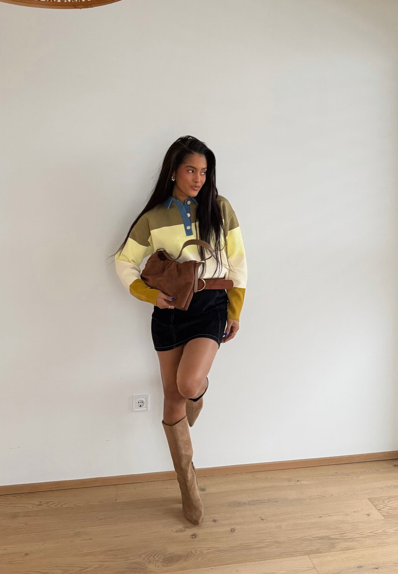 Donna con maglione a blocchi di colore, mini gonna nera, stivali marroni al ginocchio, che tiene una borsa marrone, in piedi contro una parete bianca su pavimento di legno.