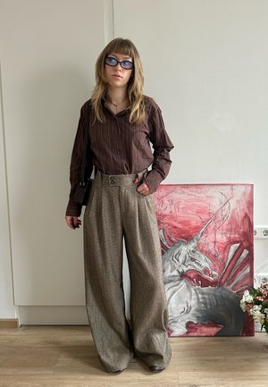 Joven con gafas junto a una detallada pintura de un unicornio rojo y gris, vistiendo una camiseta marrón a rayas y pantalones anchos beige.