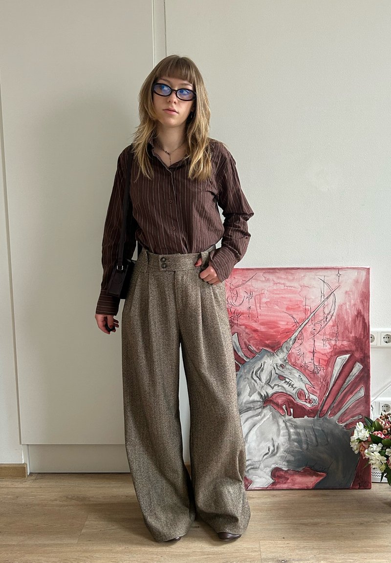 Persoană tânără cu ochelari stă lângă o pictură detaliată de unicorn roșu și gri, purtând un tricou maro cu dungi și pantaloni largi bej.