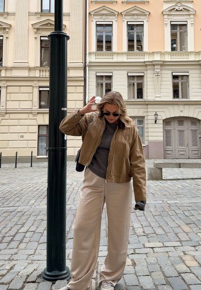 Chaqueta bomber de cuero marrón sobre un suéter gris, combinada con pantalones de pierna ancha beige claros y zapatillas blancas, en un entorno de calle adoquinada.
