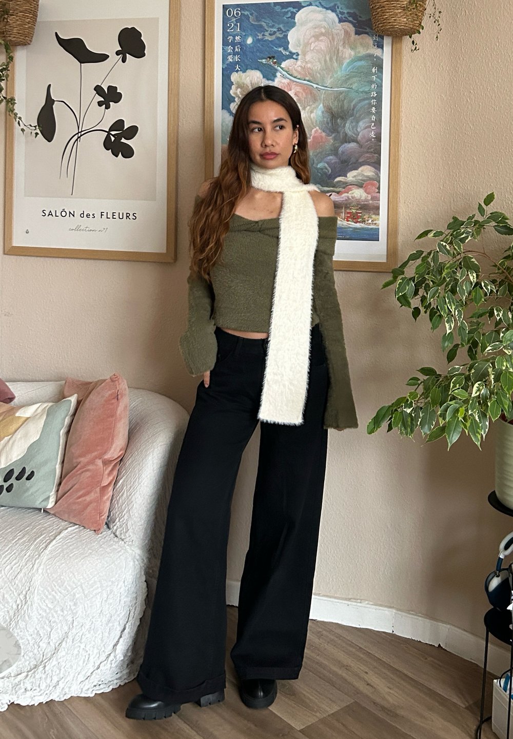 Grüner schulterfreier Pullover, weißer fluffiger Schal, schwarze weit geschnittene Hose und klobige schwarze Stiefel, vor einem neutralen Hintergrund mit Pflanzen.