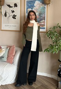Grüner schulterfreier Pullover, weißer fluffiger Schal, schwarze weit geschnittene Hose und klobige schwarze Stiefel, vor einem neutralen Hintergrund mit Pflanzen.
