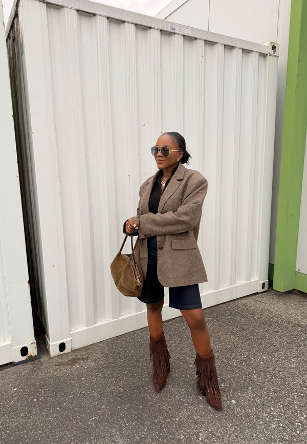 Woman with sunglasses wearing a brown blazer, black top, navy shorts, fringed brown boots, holding a brown suede handbag, standing on asphalt against a white container.