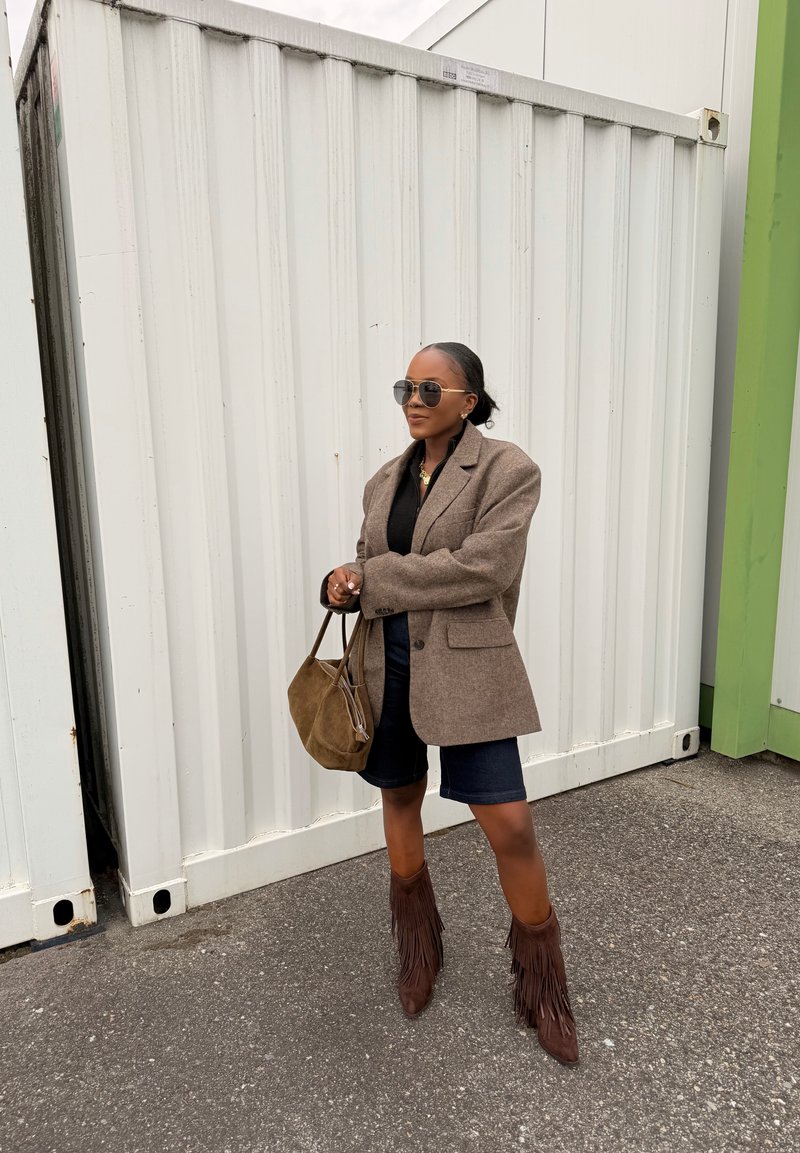 Donna con occhiali da sole che indossa un blazer marrone, top nero, pantaloncini blu scuro, stivali marroni con frange, che tiene una borsa in camoscio marrone, in piedi sull'asfalto davanti a un container bianco.