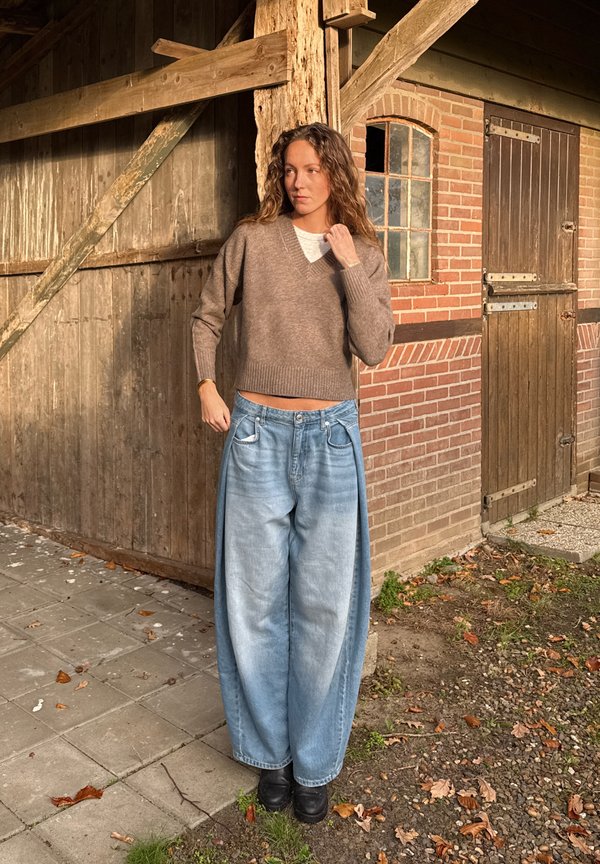 Brun stickad tröja med V-ringning, kombinerad med oversized ljusblå jeans. Mörka skor och en rustik utomhusmiljö syns i bakgrunden.