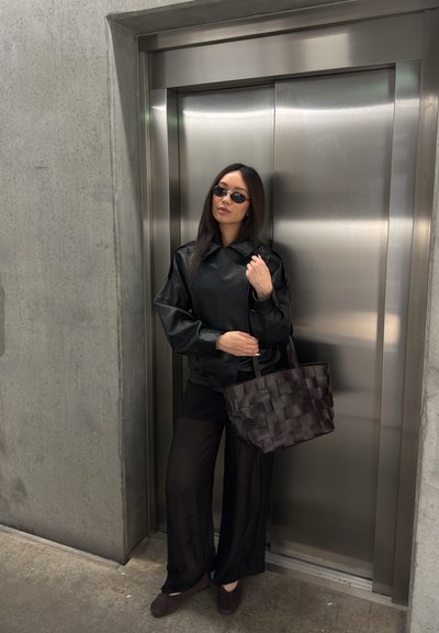 Mujer con chaqueta de cuero negra, pantalones negros transparentes y gafas de sol, está de pie sosteniendo un bolso tejido frente a unas puertas metálicas cerradas de ascensor.