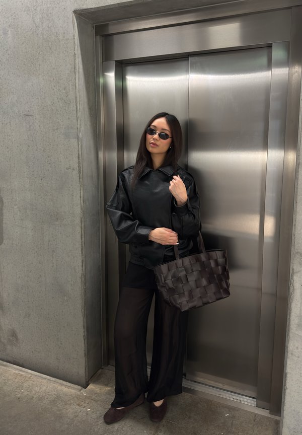 Frau in schwarzer Lederjacke, durchsichtiger schwarzer Hose und Sonnenbrille steht mit einer gewebten Handtasche vor geschlossenen Metall-Aufzugstüren.
