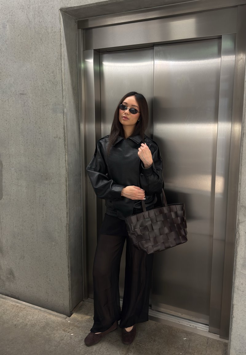 Woman in black leather jacket, sheer black pants, and sunglasses stands holding woven handbag in front of closed metal elevator doors.