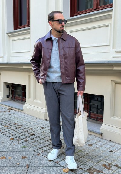 Hombre con gafas de sol, chaqueta de cuero marrón, suéter gris y pantalones, sosteniendo una bolsa de mano beige, de pie en una acera pavimentada junto a un edificio.