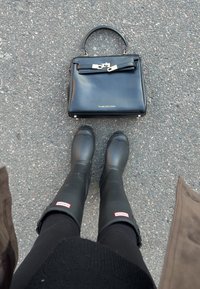 Svart skinnveske med gullfarget hardware og sløyfedetalj, plassert på asfalt ved siden av svarte gummistøvler. Føtter i svarte leggings er synlige.